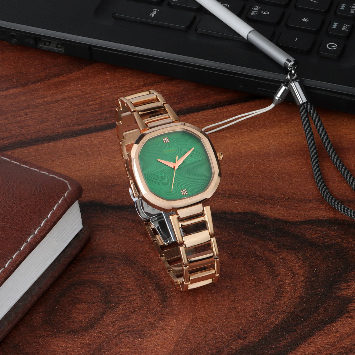 Elegant analog wristwatch with green dial and gold strap on wooden desk near notebook and laptop