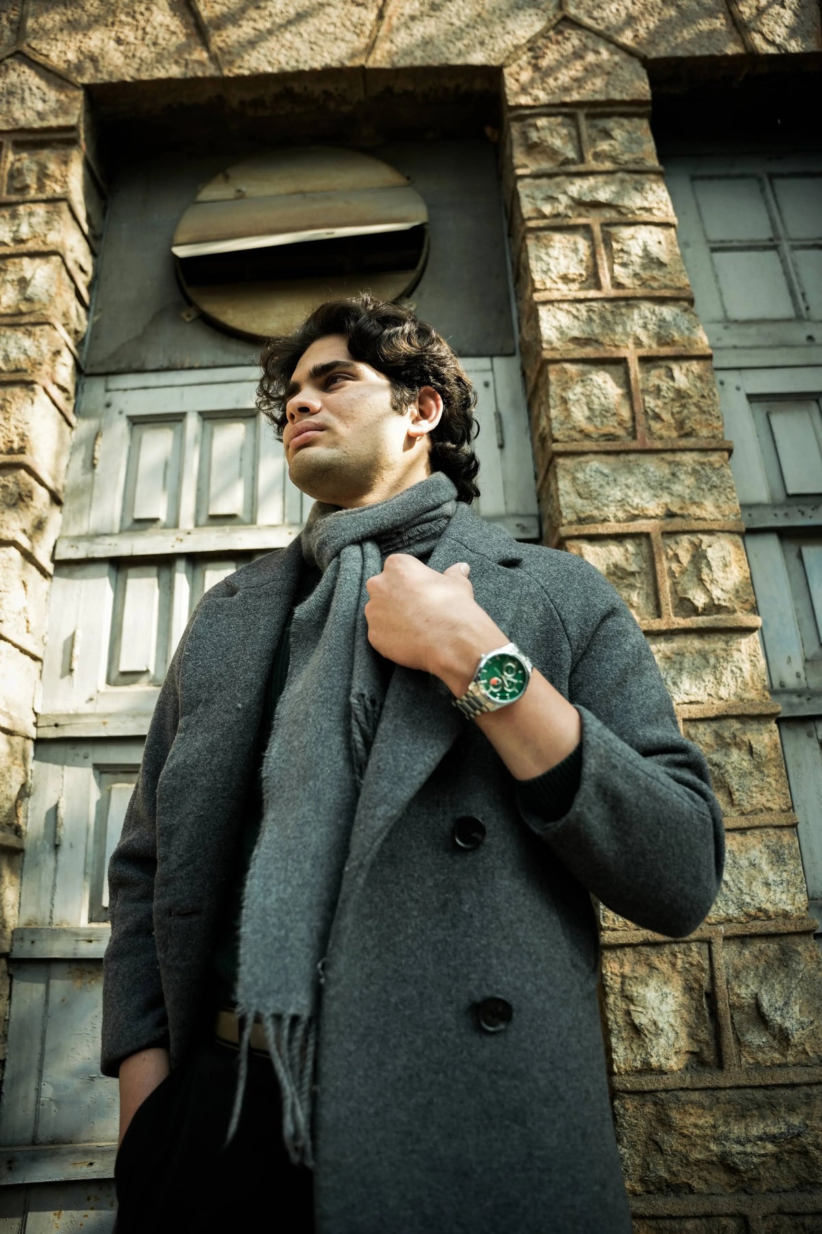 Man in grey coat wearing green dial analog quartz watch, standing by stone building