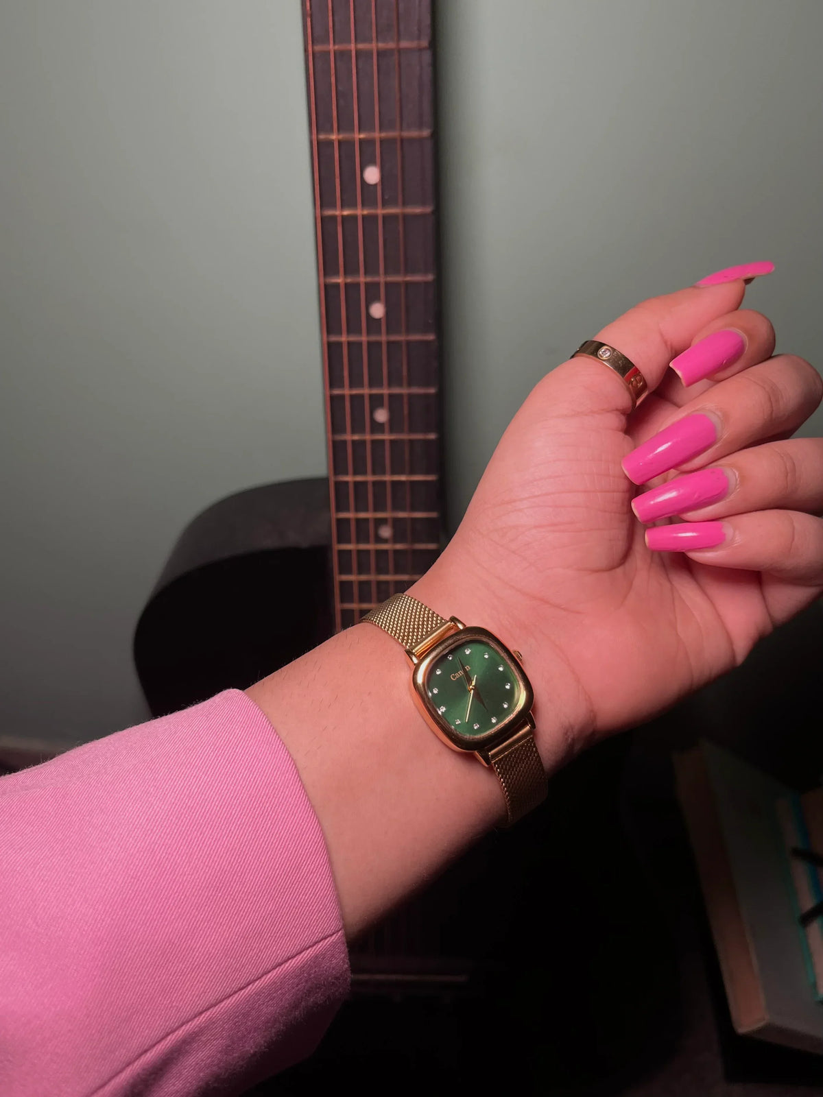 Gold analog women's watch with green dial on wrist, pink nails, guitar background