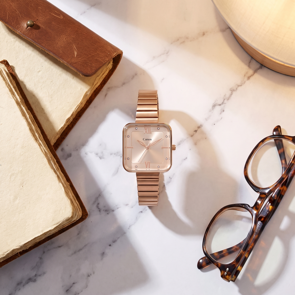 Rose gold square analog watch with Roman numerals on marble, beside leather journal and tortoise glasses