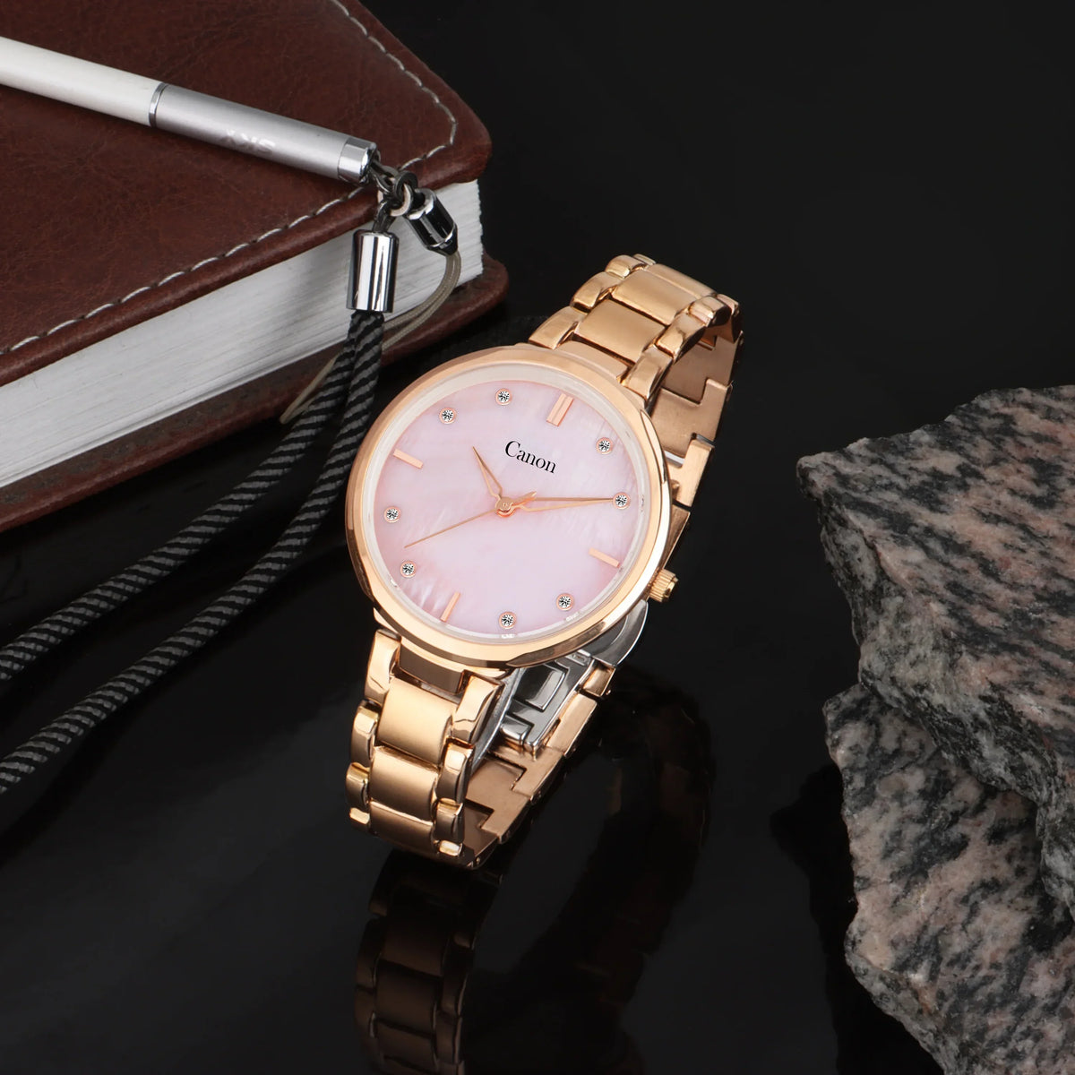 Rose gold analog watch with pink dial on metal band, displayed beside a notebook and stone