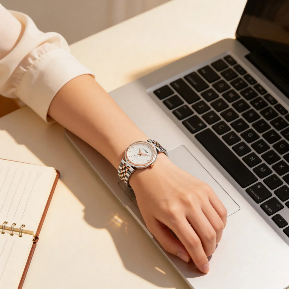 Woman wearing a silver analog wristwatch typing on a laptop at a desk with a notebook