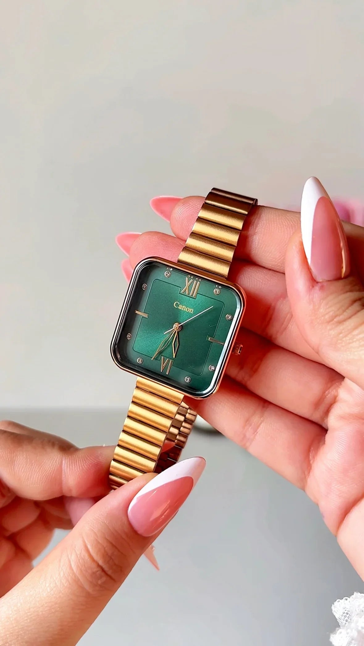 Hand holding gold metal band wristwatch with square emerald green dial and Roman numerals