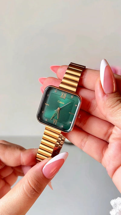 Hand holding gold metal band wristwatch with square emerald green dial and Roman numerals