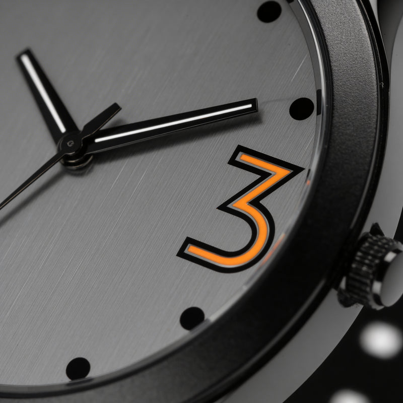 Close-up of an analog watch face with black hands, brushed silver dial, and bold orange number 3.
