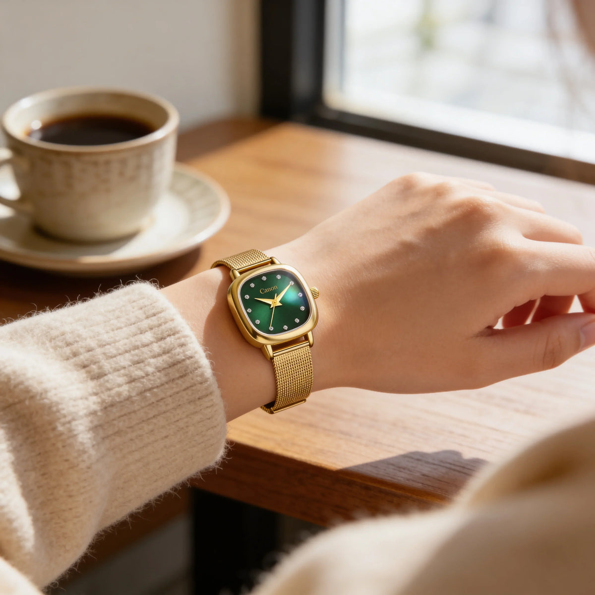 Elegant gold watch with green dial and diamond markers on wrist, beside coffee cup near window