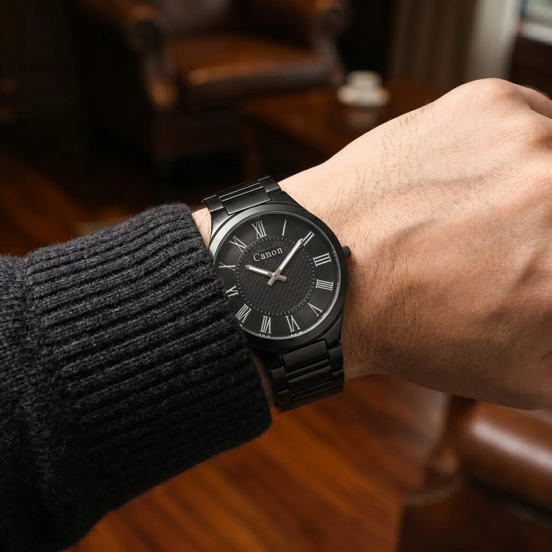 Men's black analog watch with Roman numerals and metal band, worn on wrist with sweater sleeve.
